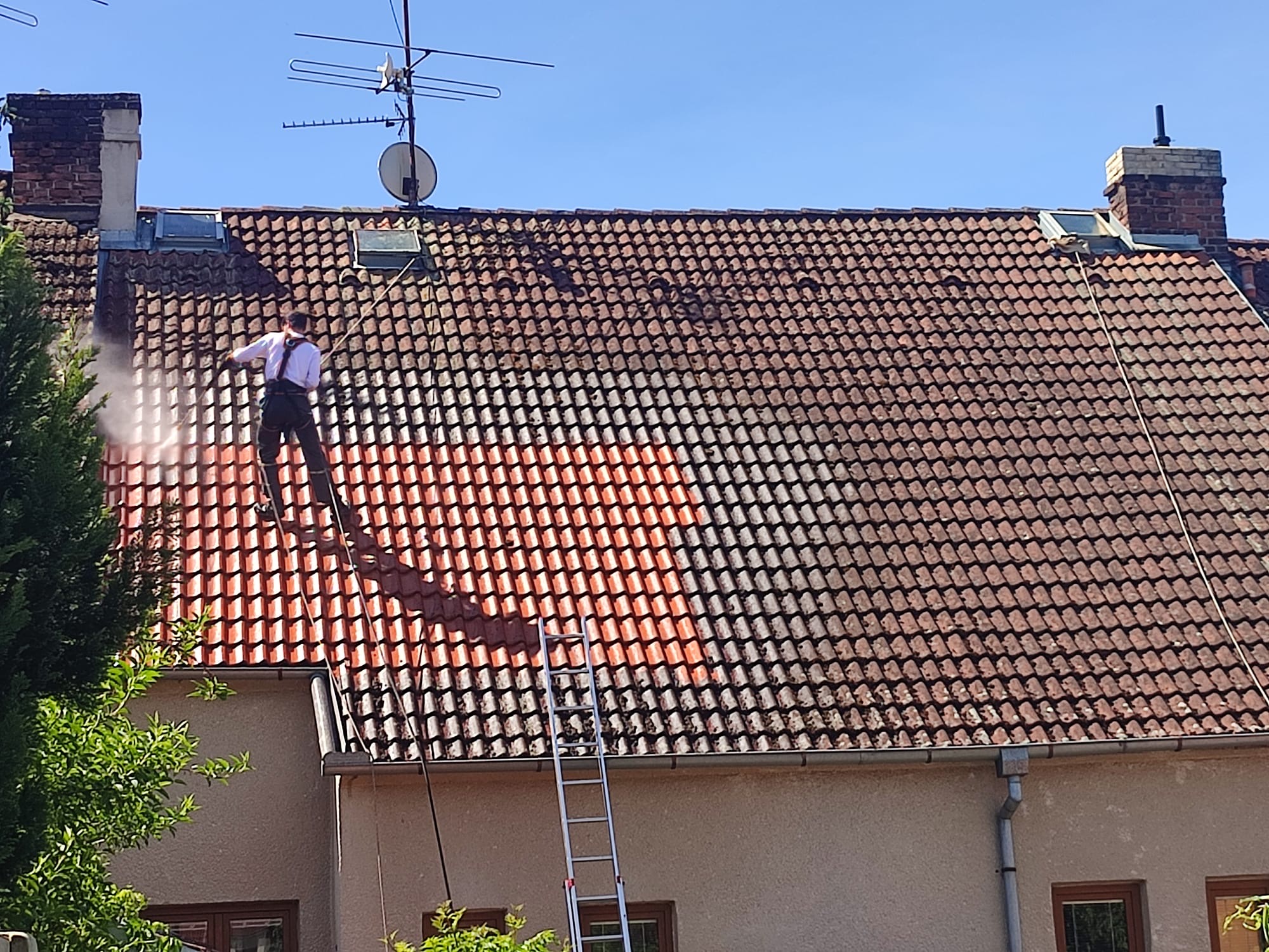 Jištěný pracovník na šikmé střeše odstraňuje mech a lišejníky z taškové krytiny