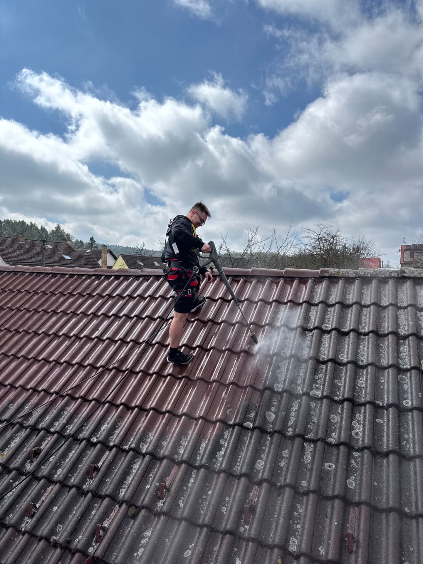 Pracovník zajištěný lanem myje znečištěnou taškovou střechu metodou Soft Wash